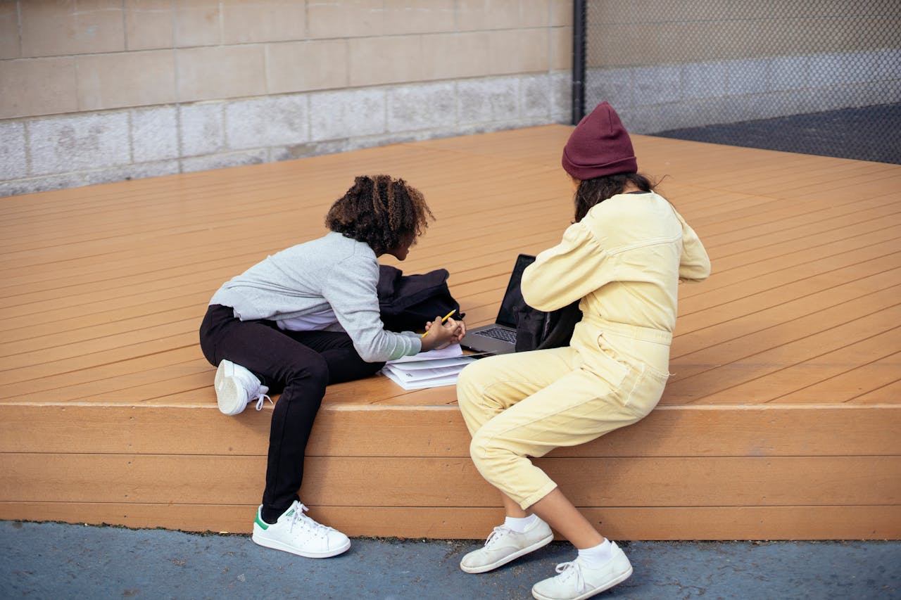Two girls studying outdoors on a wooden platform, using a laptop together.